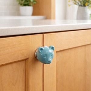 May include: A light blue ceramic cabinet knob in the shape of a bear's head. The knob is mounted on a light wood cabinet door. The background includes a white countertop, a small potted plant, and a vase of yellow flowers.