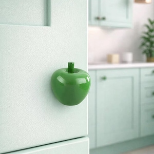 Könnte beinhalten: Ein glänzender, grüner, apfelförmiger Schrankknauf ist an einer hellgrünen Schranktür befestigt. Der Knauf hat einen Stiel und einen abgerundeten Körper. Der Hintergrund zeigt eine Küche mit passenden grünen Schränken.