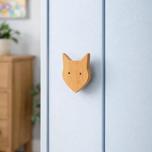 Könnte beinhalten: Ein schrankknopf in Form eines Fuchses aus Holz. Der Knopf ist hellbraun mit zwei kleinen, runden Augenlöchern. Er ist an einer hellblauen Schranktür befestigt. Der Knopf ist ein dekoratives Element für Kindermöbel.
