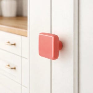 以下が含まれることがあります： 丸みを帯びたエッジを持つ正方形のコーラル色のキャビネットノブが、白いキャビネットドアに取り付けられています。ノブには小さな円形のベースがあります。背景には、金色のハードウェアを備えた他の白いキャビネットが見えます。