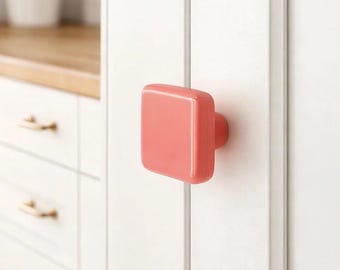 Può includere: Un pomello quadrato, color corallo, con un bordo arrotondato, è montato su un'anta bianca. Il pomello ha una piccola base circolare. Altri mobili bianchi con ferramenta dorata sono visibili sullo sfondo.