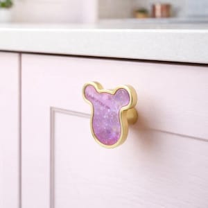May include: A decorative cabinet knob. It features a gold-coloured frame and a purple, speckled centre. The knob is attached to a light pink cabinet door, adding a touch of elegance.