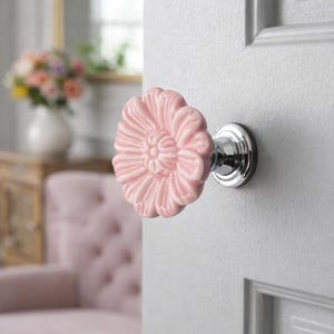 May include: A pink ceramic flower-shaped door knob with a chrome base is attached to a white door. The knob has detailed petal designs. The background shows a glimpse of a room with a pink sofa and a vase of flowers.