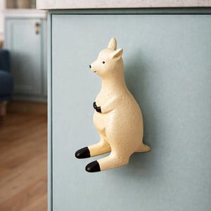 May include: A cream kangaroo-shaped cabinet knob with black details, mounted on a light blue cabinet door. The kangaroo is depicted in a sitting pose with crossed arms. The knob has a textured surface.