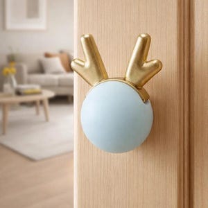 Puede incluir: Un pomo de puerta azul claro y dorado con un diseño de cuernos de ciervo. El pomo es redondo con cuernos dorados y está montado en una puerta de madera clara. El fondo muestra un salón borroso.