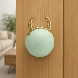 Può includere: Un pomello decorativo per porta con un centro sferico verde chiaro e corna color oro. Il pomello è montato su una porta di legno marrone chiaro. Il design ricorda una testa di toro, aggiungendo un tocco unico all'arredamento della casa.