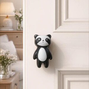 Op de afbeelding: Een zwart-witte deurknop in de vorm van een panda. De knop is bevestigd op een witte deur. De panda heeft een wit gezicht en buik, met zwarte accenten rond de ogen, oren en ledematen. De deur heeft een verzonken paneelontwerp.