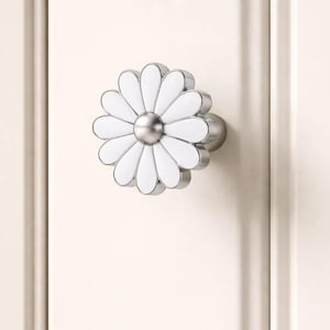 May include: A daisy-shaped cabinet knob with a white enamel finish and a brushed nickel center and base. The floral design adds a decorative touch to furniture. The knob is mounted on a light-colored cabinet door.