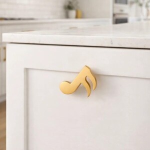 May include: A gold-colored cabinet pull in the shape of a musical eighth note. The hardware is mounted on a white cabinet door in a kitchen setting. The kitchen has white cabinets and a white countertop.