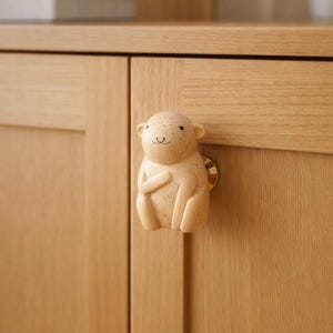 May include: A light-coloured wooden cabinet knob shaped like a seated animal with a smiling face. The knob is attached to a light-coloured wooden cabinet door with a brass-coloured screw. The cabinet is made of light-coloured wood.