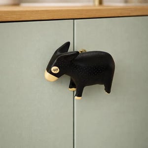 Peut inclure: Bouton de meuble noir en forme d'âne avec des détails dorés. Le bouton est fixé à une porte d'armoire vert clair. L'âne a une texture pointillée et un design fantaisiste, idéal pour la décoration.