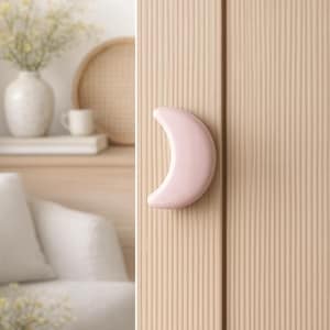 Op de afbeelding: Een lichtroze, halvemaanvormige kastgreep. De greep is bevestigd aan een lichtbeige, verticaal gegroefde kastdeur. De deur maakt deel uit van een neutraal interieurontwerp.