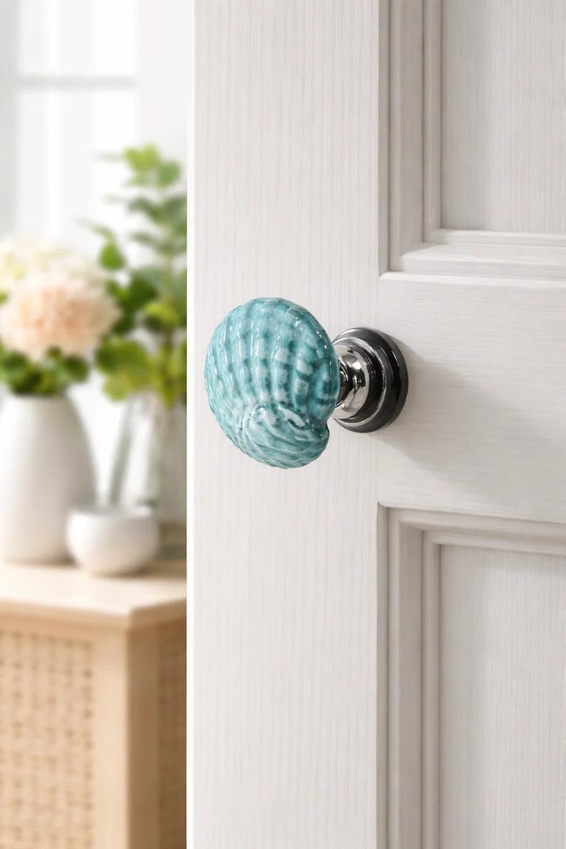 Może przedstawiać: Gałka do drzwi w kształcie muszli w odcieniach turkusu i bieli, zamontowana na białych drzwiach. Gałka ma chromowaną podstawę i czarną płytkę tylną. Drzwi są drewniane.