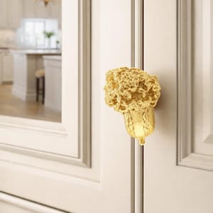May include: A gold-colored cabinet door pull with a unique, textured design. The pull is shaped like a stylized flower or organic form, adding a decorative element to the white cabinet doors. The background shows a glimpse of a kitchen.