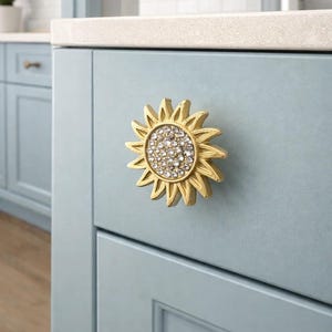 Może przedstawiać: Złoty uchwyt do szafki w kształcie słonecznika z wysadzanym kryształami środkiem jest zamontowany na jasnoniebieskich drzwiach szafki. Wzór słonecznika ma szczegółowe płatki. Uchwyt dodaje dekoracyjnego akcentu.