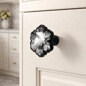 May include: A clear, faceted crystal cabinet knob with a black, floral-shaped base. The knob is mounted on a white cabinet door, offering a touch of elegance to kitchen or bathroom decor.