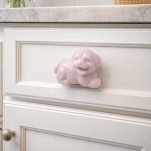 以下が含まれることがあります： 薄いピンク色のセラミック製の子犬型キャビネットノブ。ノブは白いキャビネットのドアに取り付けられています。子犬は笑顔で横たわっています。キャビネットはキッチンの設定です。