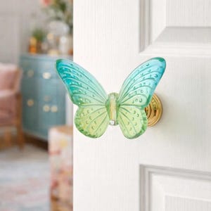 May include: A butterfly-shaped door knob. The knob is made of translucent glass, with a green to blue gradient. It is mounted on a gold-coloured base and attached to a white door. The door knob is a decorative item.