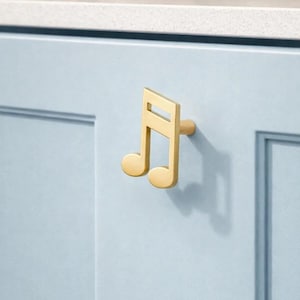 Może przedstawiać: Złoty uchwyt do szafki w kształcie ósemki nutowej. Uchwyt jest zamontowany na jasnoniebieskich drzwiach szafki. Design jest nowoczesny i minimalistyczny, odpowiedni do kuchni lub łazienki. Wymiary w centymetrach.