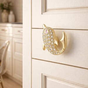Könnte beinhalten: Goldfarbener Schrankknauf in Delfinform, besetzt mit klaren Kristallen. Der Knauf ist an einer cremefarbenen Schranktür befestigt. Das Design des Delfins verleiht Küchen- oder Badmöbeln eine dekorative Note.