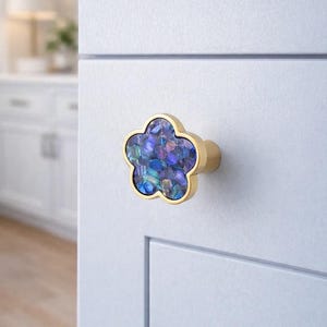 May include: A floral cabinet knob with a gold finish and a centre filled with iridescent blue and purple pieces. The knob is mounted on a white cabinet door, a decorative hardware element.