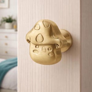 Op de afbeelding: Een messing paddenstoelvormige deurknop. De knop is bevestigd op een lichtgekleurde houten deur. De paddenstoelkap heeft cirkelvormige inkepingen en de steel heeft een afgeronde vorm. Een decoratief item voor een deur.