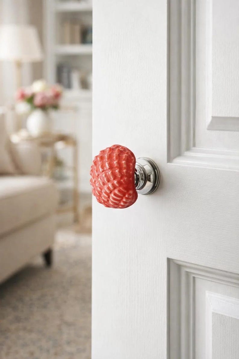 Może przedstawiać: Czerwona, teksturowana gałka do drzwi z motywem muszli. Gałka jest zamontowana na białych drzwiach ze srebrną podstawą. Tło jest rozmyte, ukazując fragment salonu.