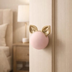 Può includere: Un pomello per porta rosa e oro a forma di muso di maiale. Il pomello ha un corpo rotondo rosa con orecchie dorate. È montato su una porta di legno colorata.