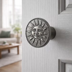 Puede incluir: Un pomo de puerta plateado con un diseño de sol. El pomo redondo presenta una cara de sol detallada en el centro, rodeada de rayos radiantes y un borde de cuentas. El pomo está montado en una puerta blanca, lo que sugiere un elemento de decoración del hogar.