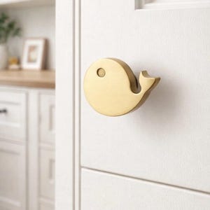 May include: A brass whale-shaped cabinet knob mounted on a white door. The knob has a circular body with a small eye and a curved tail fin. The background shows a glimpse of a kitchen with white cabinets and a wooden countertop.