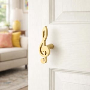 Könnte beinhalten: Ein goldfarbener Türgriff in Form eines Notenschlüssels ist an einer weißen Tür angebracht. Der Griff hat einen zylindrischen Sockel und ein dekoratives Musiksymbol. Die Tür befindet sich in einem Raum mit unscharfem Hintergrund.