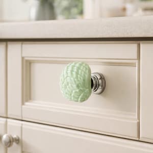 Op de afbeelding: Lichtgroene keramische kastknop met een textuur, schelpachtig ontwerp. De knop is gemonteerd op een chromen basis en bevestigd aan een crèmekleurige kastdeur. De keukenomgeving omvat een aanrecht en een vaas met bloemen.