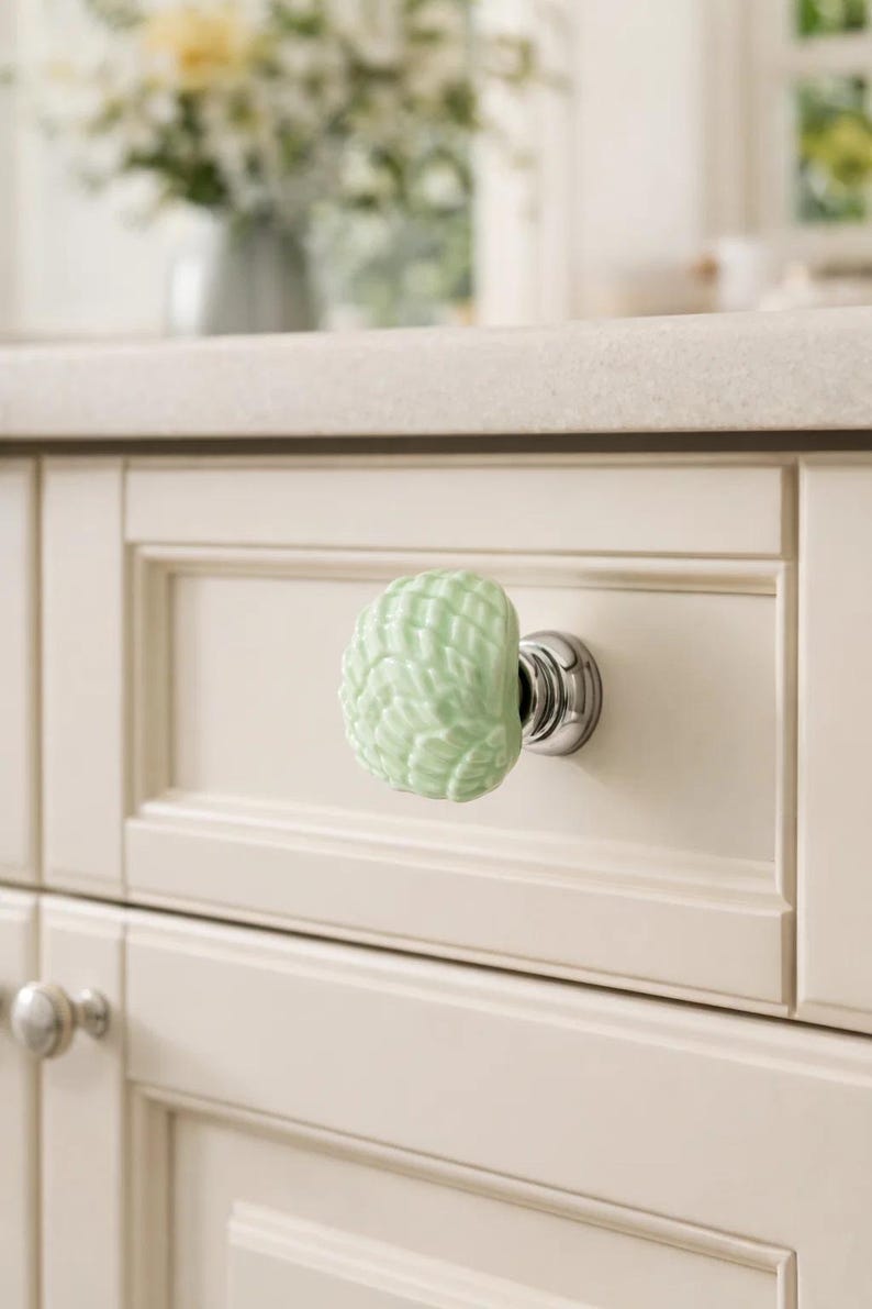 Może przedstawiać: Jasnozielony ceramiczny uchwyt do szafki z teksturowanym, przypominającym muszlę wzorem. Uchwyt jest zamontowany na chromowanej podstawie i przymocowany do kremowych drzwi szafki. Otoczenie kuchni obejmuje blat i wazon z kwiatami.