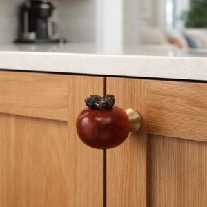 Może przedstawiać: Zbliżenie drzwi szafki kuchennej z unikalnym, okrągłym, czerwonobrązowym uchwytem. Uchwyt ma ciemną, teksturowaną górę i mosiężną podstawę. Szafka wykonana jest z jasnego drewna.