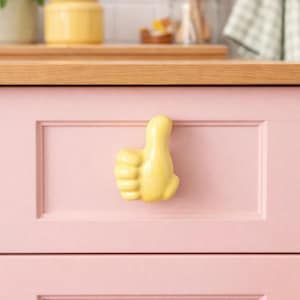 May include: A yellow thumbs-up shaped cabinet pull is mounted on a pink cabinet door. The kitchen setting includes a wooden countertop, a yellow jar, and a checkered towel.