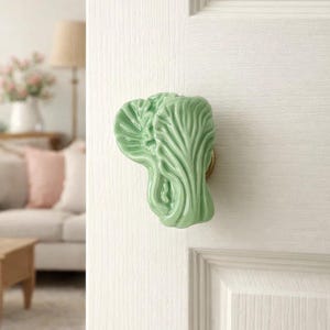 May include: A light green ceramic door knob shaped like a head of lettuce. The knob is mounted on a white door with a gold-colored base. The door is part of a room with a couch and lamp.