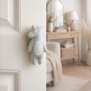 Pode incluir: Um puxador de porta de cerâmica cinza em forma de urso de desenho animado, com barriga e rosto brancos, fixado a uma porta branca. O puxador em forma de urso adiciona um toque lúdico ao design da porta. O puxador é um elemento decorativo.