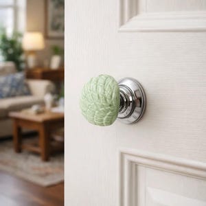 Może przedstawiać: Jasnozielona, ​​teksturowana ceramiczna gałka do drzwi z podstawą z metalu w kolorze srebrnym jest przymocowana do białych drzwi. Gałka ma zaokrąglony kształt ze szczeg&oacute;łowym, liściastym wzorem. Drzwi są drewniane.