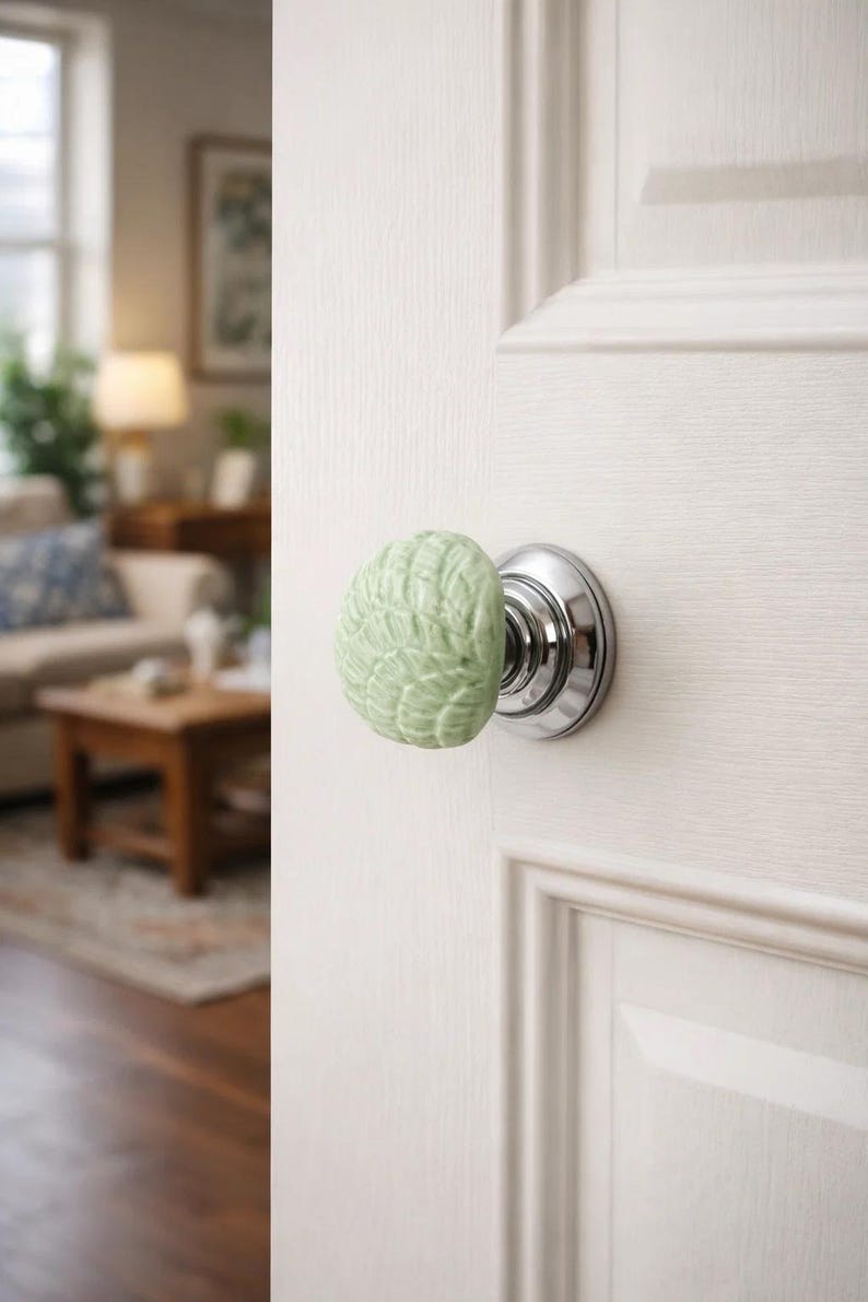 Może przedstawiać: Jasnozielona, ​​teksturowana ceramiczna gałka do drzwi z podstawą z metalu w kolorze srebrnym jest przymocowana do białych drzwi. Gałka ma zaokrąglony kształt ze szczeg&oacute;łowym, liściastym wzorem. Drzwi są drewniane.