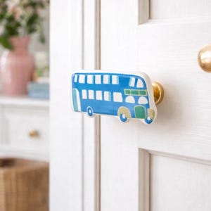 Könnte beinhalten: Blau-weißer Doppeldeckerbus-Türknauf mit goldenen Beschlägen. Das Busdesign zeigt Fenster und Räder mit grünen und gelben Akzenten. Der Knauf ist an einer weißen Tür befestigt, ein dekoratives Wohnaccessoire.