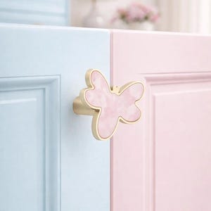 Könnte beinhalten: Ein schmetterlingsförmiger Schrankknauf mit goldener Oberfläche und einem rosa marmorierten Mittelteil. Der Knauf ist an einer hellblauen und rosa Schranktür angebracht. Der Knauf verleiht Möbeln eine dekorative Note.