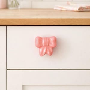 May include: A pink bow-shaped cabinet knob is fixed to a white drawer. The glossy, decorative knob is a charming accent. The drawer is part of a white cabinet with a wooden countertop in the background.