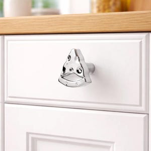 Puede incluir: Un tirador de armario plateado brillante con un diseño triangular único. El tirador está montado en una puerta de armario blanca, con una encimera de madera visible en el fondo. El tirador tiene una forma moderna y geométrica.