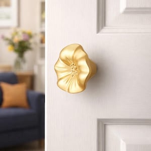 May include: A gold flower-shaped door knob fixed to a white door. The knob features a detailed floral design with a textured surface and a central cluster of small, round details. The door has a panel design.