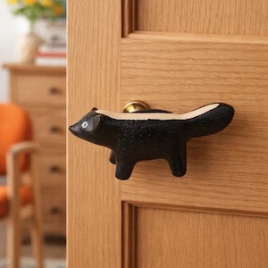 May include: A black and white skunk-shaped door knob. The skunk has a white stripe down its back and is mounted on a light brown wooden door. The door knob is made of a solid material and has a brass-colored base.