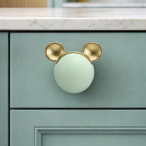 May include: A mint green cabinet knob with gold-colored ears, resembling a cartoon character. The knob is mounted on a light blue cabinet door. The design adds a playful touch to kitchen or bathroom cabinetry.