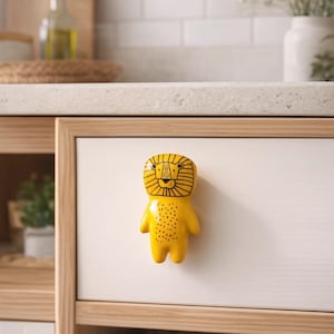 Könnte beinhalten: Ein gelber, löwenförmiger Schrankknauf mit schwarzen Details ist an einer weißen Schranktür befestigt. Der Knauf hat ein cartoonartiges Design mit einem lächelnden Gesicht und kleinen schwarzen Punkten. Der Schrank ist in einem hellen Holzrahmen.