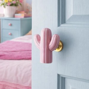 Può includere: Un pomello per porta a forma di cactus in ceramica rosa con base dorata è fissato a una porta azzurra. La porta fa parte di una camera da letto con un letto coperto da biancheria da letto rosa e un comodino blu.
