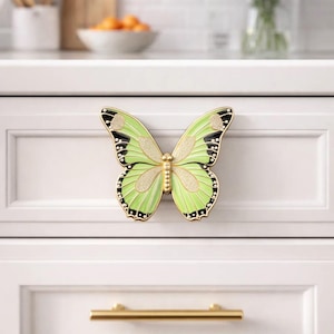 Peut inclure: Un bouton de meuble décoratif en forme de papillon, vert clair et noir. Le papillon a des accents de paillettes dorées et est monté sur une porte d'armoire blanche. Une poignée dorée est en dessous.