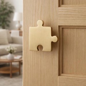 May include: A gold-colored puzzle piece door handle mounted on a light-colored wooden door. The handle is a single piece with a square shape and rounded edges, and is a decorative element.